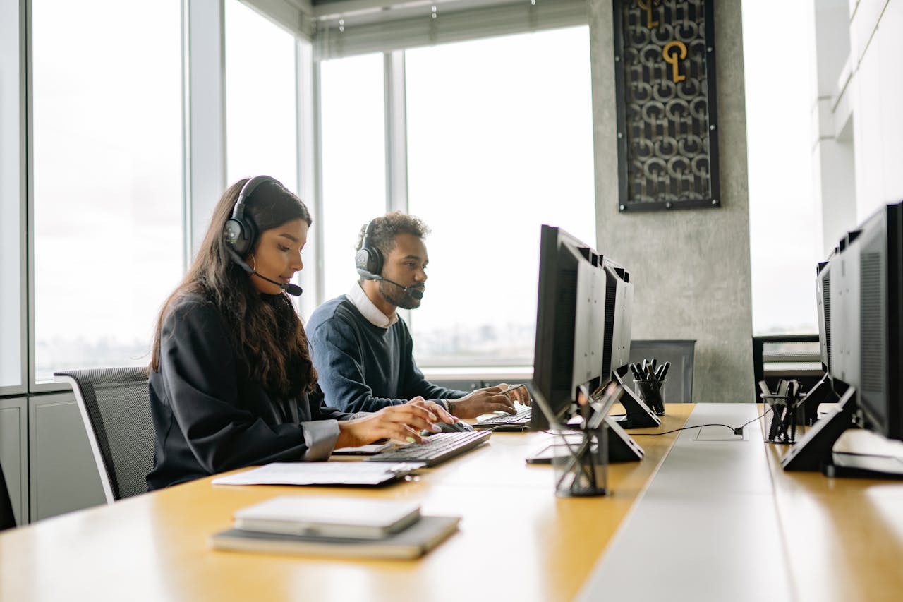 About Two customer service representatives wearing headsets working at computers in a modern office.